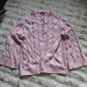 Ljusrosa kabelstickad kofta Gina Tricot - Supersöt ljusrosa kofta från Gina Tricot med grov kabelstickning och pärlemorknappar framtill. Koftan har ribbade muddar och rund hals, perfekt för lager på lager under höst och vinter. Mysig och trendig att slänga på sig när det är kyligt.