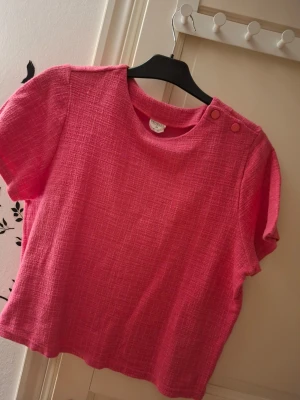 Rosa strukturerad topp med knappar - En rosa topp från Cubus i strukturerat tyg med korta ärmar och rund hals. Två dekorativa knappar på ena axeln ger en snygg detalj. Passformen är normal och materialet känns mjukt och bekvämt mot huden.