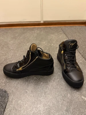 Svarta high-top sneakers med gulddragkedjor - Säljer ett par svarta high-top sneakers från Giuseppe Zanotti med guldfärgade dragkedjor på sidorna och svart snörning. Skorna är tillverkade i skinn och har en rund tå samt svart sula. Insidan är beige och plösen pryds av den ikoniska guldfärgade loggan.