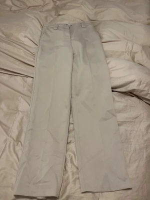 Beige kostymbyxor från NA-KD - Säljer ett par stilrena beige kostymbyxor från NA-KD i storlek 34. Byxorna har raka ben, bälteshällor och två fickor bak. Materialet är mjukt och har en lätt struktur, perfekt för en clean och snygg look.