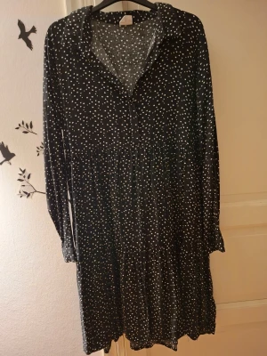 Svart prickig långärmad klänning - Svart klänning med vita prickar och långa ärmar. Klänningen har en lös passform, krage och knappar framtill. Tyget känns mjukt och faller snyggt, perfekt för dig som gillar en avslappnad men ändå stilsäker look.