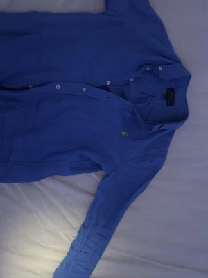 Blå skjorta från Polo Ralph Lauren - Klassisk blå skjorta från Polo Ralph Lauren med button-down krage och vita knappar. Ikoniska gula loggan broderad på bröstet. Tillverkad i mjuk bomull och har lång ärm. Perfekt för en clean och stilren look.
