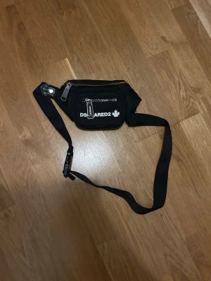 Svart magväska från Dsquared2 - Säljer en svart magväska från Dsquared2 med justerbart band och två dragkedjefack. Väskan har Dsquared2-logga och ett vitt lönnlöv på framsidan. Perfekt för att bära det viktigaste och enkel att slänga över axeln eller runt midjan.