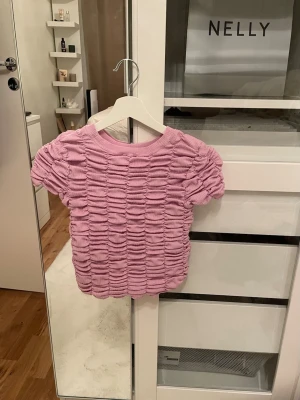 Ljuslila ribbad topp med puffärm - Superfin ljuslila topp från by ANTHROPOLOGIE  med korta puffärmar och ett ribbat, rynkat tyg som ger en cool struktur. Toppen har rund halsringning och är croppad i modellen. Perfekt att styla med jeans eller kjol för en trendig look. (OBS! köpt utomlands och det är en liten tråd som åkt ut)