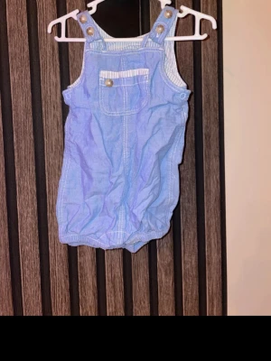 Blå byxdress med knappar, storlek 62 - Söt blå byxdress i bomull med justerbara axelband och dekorativa knappar framtill. Har en bröstficka med randig kant och resår vid bensluten för en bekväm passform. Perfekt för varma dagar. Storlek 62