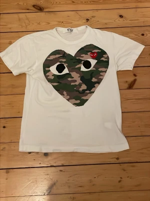 Vit Comme des Garçons Play t-shirt - Säljer en vit t-shirt från Comme des Garçons Play i storlek M. Framsidan har ett stort hjärta i camouflage med ögon och en liten röd hjärtdetalj med ögon. Klassisk passform och mjuk bomullskänsla. Perfekt för dig som gillar streetwear och statement-plagg. Kan diskutera priset också👍