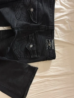 Svarta True Religion jeans bootcut - Säljer ett par svarta jeans från True Religion med ikoniska ficklock och silverfärgade knappar. Bootcut-modell med klassisk label bak och snygga sömdetaljer. Jeansen är i jeansmaterial och har dragkedja framtill. Sitter jätteskönt!!!