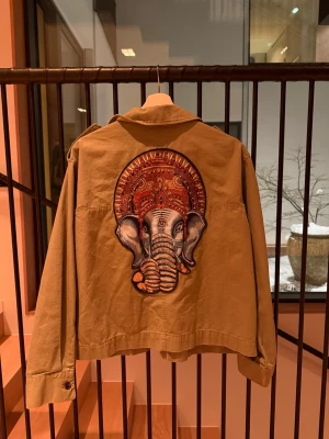 Grön jacka från H&M med elefantbrodyr - Säljer en beige overshirt från H&M i bomull med stor broderad elefant på ryggen. Jackan har två stora fickor framtill, axelklaffar och knappar vid ärmslut. 