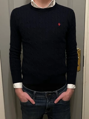 Mörkblå stickad tröja från Morris - Tjena! 🍾 Säljer en mörkblå stickad tröja från Morris med klassiskt kabelstickat mönster och rund hals. Tröjan har en liten röd logga broderad på bröstet och långa ärmar. Perfekt att slänga på över en skjorta för en clean look. Tröjan är liten i storleken som motsvarar Xs. Ifall du är tveksam över något, tveka inte fråga på! ✨