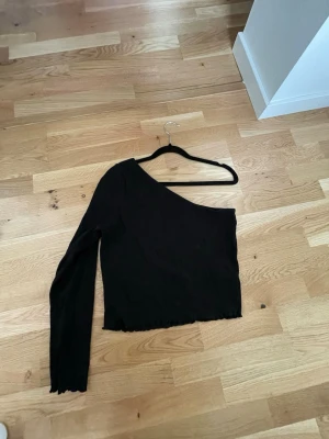 Svart oneshoulder topp - En svart oneshoulder ribbad topp med lång ärm på ena sidan. Toppen är lite croppad och har vågig kant nedtill för en extra detalj. Oanvänd!