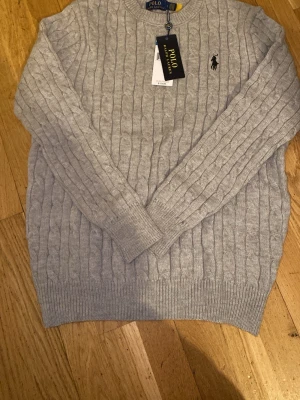 Grå kabelstickad tröja Polo Ralph Lauren - Kabelstickad tröja från Polo Ralph Lauren i ljusgrått. Klassisk rund halsringning, ribbade muddar och ikoniska broderade loggan på bröstet. Perfekt för lager på lager under höst och vinter. Skön och stilren, passar till allt. Aldrig använd. 