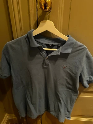 Blå pikétröja från Gant - Klassisk blå pikétröja från Gant med korta ärmar och krage. Tröjan har två knappar framtill och en liten broderad logga på bröstet. Tillverkad i bomull för en skön och luftig känsla.