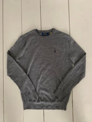 Grå merinoulltröja Polo Ralph Lauren - Snygg grå långärmad tröja från Polo Ralph Lauren i tvättbar merinoull. Slim fit-modell med rund hals och diskret logga broderad på bröstet. Perfekt för dig som gillar stilrena och klassiska plagg med skön passform.