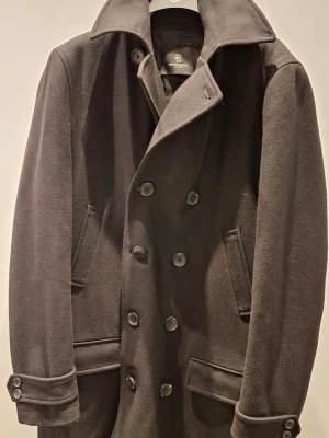 Svart skepparkavaj från Bruuns Bazaar - Superfin dubbelknäppt kashmirrock från Bruuns Bazaar, herr i storlek Medium. Två bröstfickor samt två nedre fickor. Quiltat innerfoder med två innerfickor. Mycket bra skick då den inte kommit till användning. Pris; 1000 kr (nypris 4999 kr)