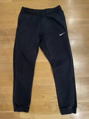 Svarta mjukisbyxor från Nike, stl S - Svarta mjukisbyxor från Nike med klassisk vit swoosh-logga på benet. Byxorna har elastisk midja och muddar vid bensluten för en skön passform. Tillverkade i mjukt bomullsmaterial som är perfekt för chill eller träning.
