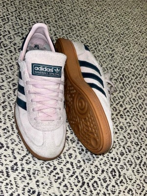Adidas Handball Spezial ljusrosa sneakers - Adidas Handball Spezial sneakers i ljusrosa mocka med mörkblå ränder och detaljer. Skorna har klassisk gummisula, snörning och rund tå. Insidan är ljusrosa med Adidas-logga. Perfekt för dig som gillar retrostil och snygga färgkombinationer. Nypris ca 1250kr! 
