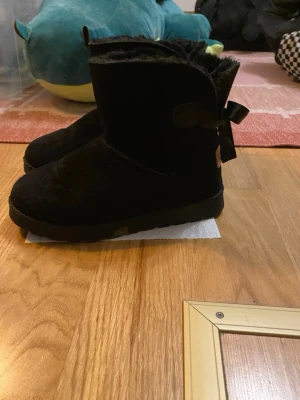 Svarta boots med rosett från REX - Mysiga svarta boots från REX med fluffigt foder och dekorativa rosetter baktill. Skorna är i mocka och har en platt sula, perfekt för kalla dagar. Klassisk rund tå och enkel design som passar till många outfits. Säljer då de är för små, pris kan diskuteras