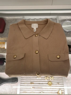 Beige kortärmad skjorta från H&M - Snygg beige skjorta från H&M i storlek M Skjortan har korta ärmar, två bröstfickor med lock och guldiga knappar framtill. Materialet är stickat och ger en mjuk känsla. Perfekt för dig som gillar stilrena detaljer och enkel design.