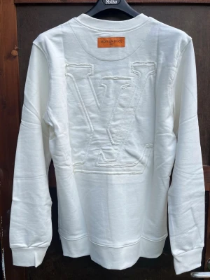 Vit LV sweatshirt XL - Säljer en stilren vit sweatshirt från Louis Vuitton i storlek XL. Tröjan har ett stort broderat LV-logo på ryggen och mindre broderade detaljer framtill. Mjuk bomullskvalitet och klassisk rund hals. Perfekt för dig som gillar exklusiva streetwear-vibbar.