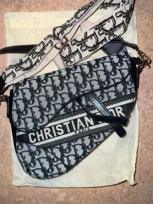 axelväska - Säljer en axelväska med svart och vit monogramdesign. Väskan har en unik sadelform, bred axelrem med logotyp och detaljer i svart skinn. Perfekt statement piece för dig som gillar lyxiga accessoarer. Pris kan diskuteras 