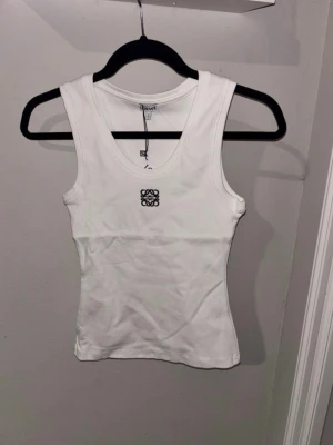 Loewe linnetopp  - Säljer en fräsch vit linnetopp från Loewe med svart broderad logga framtill. Toppen är ribbad och har en rundad halsringning samt ärmlös design. Perfekt för dig som gillar stilrena och lyxiga basplagg med exklusiv känsla.