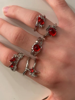 Silvriga ringar med röda stenar - Säljer ett set med fem silvriga ringar med unika former och detaljer, alla prydda med röda stenar i olika former och storlekar. Det är ej äkta silver! Styckpris 7kr, alla för 30kr! 🫶