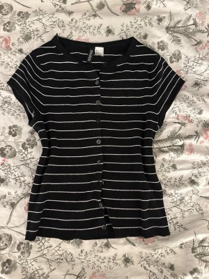 Svart och vit randig blus från Kappahl - Säljer en svart blus med vita horisontella ränder från h&m. Blusen har korta ärmar, rund halsringning och knappar hela vägen framtill. Materialet är mjukt och ribbat, perfekt för en chill look.