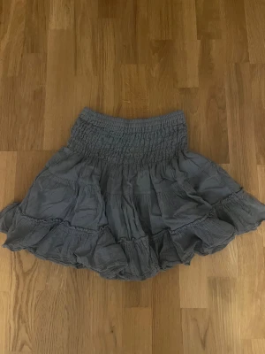 Grå volangkjol med smock - Supersöt grå kortkjol med bred smock i midjan och flera lager volanger. Köptes i en butik i Thailand och är helt oanvänd. Materialet känns mjukt och lätt mot huden.