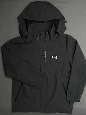 Svart vindjacka från Under Armour - Svart vindjacka från Under Armour med avtagbar huva, elastiska muddar och dragkedja framtill. Jackan har ett stilrent tryck med logga på bröstet och detaljer vid kragen. Perfekt för dig som gillar sportig och clean stil.