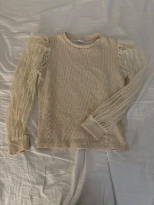 Beige tröja med spetsärmar från Zara - Supersnygg beige tröja från Zara med ribbad kropp och transparenta, puffiga spetsärmar. Ärmarna har ett vågigt mönster och muddar vid handlederna. Perfekt för dig som vill ha något unikt och trendigt i garderoben!