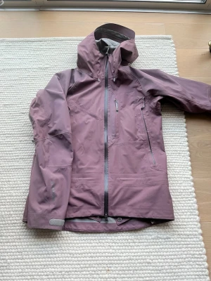 Houdini skaljacka dam M i lila - Säljer en Houdini skaljacka för dam i storlek M. Jackan är lila med huva, vattentäta dragkedjor och flera fickor framtill. Perfekt för utomhusaktiviteter och har justerbara ärmslut med kardborre. Materialet är slitstarkt och vattenavvisande syntet.