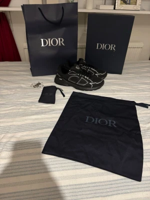 B30 - Säljer ett par svarta Dior sneakers med vita detaljer och tydlig CD-logga på sidan. Skorna har snörning, rund tå och platt sula. Materialet är en mix av mesh och syntet, vilket ger en sportig vibe. Medföljer originalpåse, box och accessoar från Dior. Allt kommer med förutom kvitto, kontakta för mer bilder 