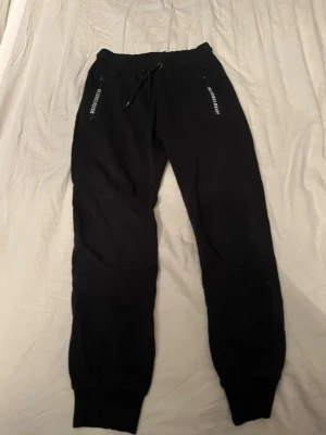 Svarta mjukisbyxor från Woxo - Svarta mjukisbyxor från Woxo Urban Streetwear med elastisk midja och snörning. Byxorna har ribbade muddar vid bensluten, dragkedjor på fickorna och en liten logga på bakfickan. Perfekta för chill eller träning.