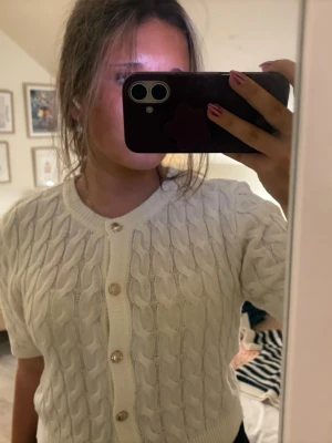 Vit kabelstickad kofta från Gina Tricot - Säljer en vit kabelstickad kofta från Gina Tricot med korta ärmar och snygga guldfärgade knappar framtill. Koftan har rund halsringning och är gjord i ett mjukt stickat material. Perfekt att slänga över en topp för en chill look.