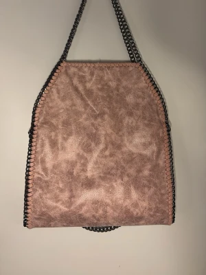 Rosa Stella McCartney liknande väska  - Säljer en oanvänd rosa axelväska med kedjehandtag och dekorativa sömmar längs kanterna. Väskan har en mjuk mockaliknande yta och dragkedja på baksidan. Insidan är ljusbeige med flera fack.