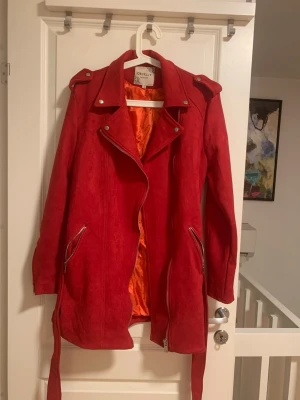 Röd mocka skinnjacka Orcelly XL - Snygg röd skinnjacka i mocka-look från Orcelly, storlek XL. Jackan har coola detaljer som dragkedjor, knappar och bälte i midjan. Fodret är orange och jackan har långa ärmar med dragkedjor vid fickorna. Perfekt för dig som vill sticka ut. Ny pris är 3000kr