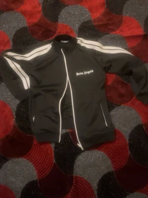 Svart zip hoodie från Palm Angels - Svart hoodie från Palm Angels med vita ränder längs ärmarna och vit dragkedja framtill. Tröjan har ståkrage, två fickor med dragkedja och logotypen tryckt på bröstet. Perfekt för en sportig och trendig look. Priset går inte att diskutera 