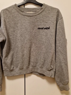 Grå sweatshirt från Pull&Bear - Säljer en grå sweatshirt från Pull&Bear med broderad text 'sweet witch' på bröstet. Tröjan har rund hals, ribbade muddar och är långärmad. Perfekt för chill dagar och enkel att matcha med jeans eller leggings.
