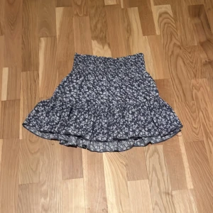 Blommig volangkjol från 157 XS - Supersöt kort kjol från 157 i storlek XS, med blå botten och vitt blommigt mönster. Kjolen har smock i midjan och volang nedtill som ger en flowy och ungdomlig vibe. Perfekt för dig som gillar att sticka ut med detaljer och mönster.