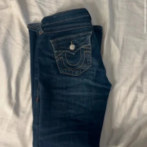 Mörkblå bootcut/lowwaist jeans true religon - Snygga mörkblå jeans från True Religion med bootcut passform. Jeansen har klassiska kontrastsömmar och ikonisk fickdetalj med lock och knapp bak. Knappt använda 1-3 gånger, vill sälja så fort som möjligt!! Kontakta innan köp! Lite förstörda längst ner men går att klippa bort/vika upp! 