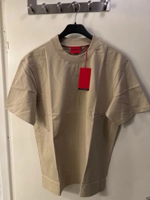Beige t-shirt från HUGO, storlek M - Snygg beige t-shirt från HUGO i mjuk bomull. Klassisk rund hals och diskret HUGO-logga på bröstet. T-shirten har korta ärmar och en clean look som funkar till många stilar. Perfekt för dig som gillar minimalistiskt och stilrent. Förpackningen är ej öppen samt ej testad.