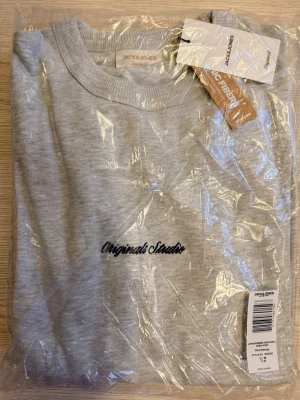 Grå sweatshirt från Jack & Jones - Snygg gråmelerad sweatshirt från Jack & Jones med broderad text 'Originals Studio' på bröstet. Tröjan har rund hals och ribbade muddar vid ärmslut och nederkant. Perfekt för chill dagar och enkel att matcha med jeans eller joggers.