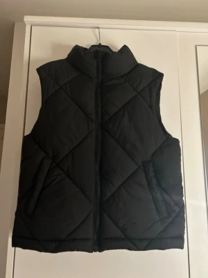 Svart väst från Gina Tricot - Säljer en svart väst från Gina Tricot i storlek 146/152. Västen har ett quiltat mönster, hög krage och dragkedja framtill. Två fickor på sidorna. Kontakta om du är intresserad!