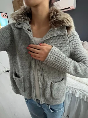 Grå stickad jacka med fuskpäls - Mysig grå stickad jacka med fluffig fuskpäls på kragen. Jackan har två framfickor, ribbade muddar och knäpps med stora knappar. Perfekt för kyliga dagar och ger en chill vibe till din outfit.