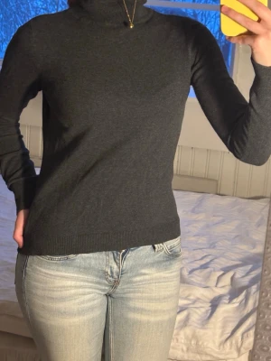 Mörkgrå polotröja i bomull - Snygg mörkgrå polotröja med lång ärm och ribbade muddar. Tröjan har en klassisk polokrage och är gjord i mjuk bomull, perfekt för lager-på-lager under kyliga dagar. Passformen är normal och den är enkel att matcha med jeans eller kjol.