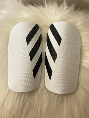 Vita Adidas benskydd med svarta ränder storlek M - Säljer ett par Adidas Tiro benskydd i storlek M, passar dig som är 140-160 cm. De är vita med svarta ränder och har en snygg, sportig design. Materialet är syntet och de kommer med en praktisk svart förvaringspåse. Perfekta för fotbollsträningen!
