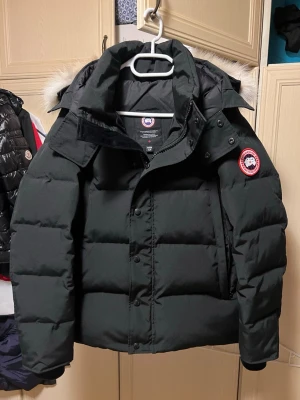 Svart pufferjacka wyndham från Canada Goose - Svart pufferjacka från Canada Goose med ikonisk röd och vit logotyp på ärmen. Jackan har huva med beige päls, quiltad design och dragkedja med knappar framtill. Perfekt för kalla vinterdagar och har en loose passform. Bra skick, har använt i 1 månad. Finns defekter, det läcker lite fjädrar i fickorna (inga hål) och jag har sytt ihop båda fickorna då stygnen gick ut. 