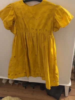 Gul klänning från Zara - Säljer en söt gul klänning från Zara med broderade blommor och håldetaljer. Klänningen har korta puffärmar och en luftig, rynkad kjol. Perfekt för dig som gillar färg och detaljer! Materialet är mjuk bomull.