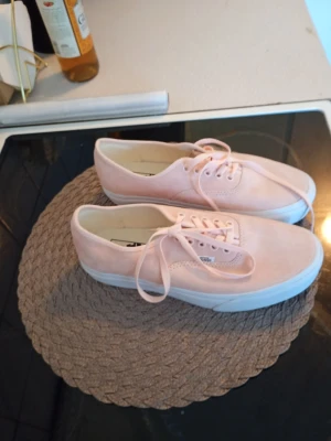 Ljusrosa Vans sneakers strl 41 - Säljer ett par snygga Vans sneakers i ljusrosa mocka med vit platt sula och rund tå. Skorna har klassisk snörning och stilren design som passar till många olika outfits. Storlek EU 41, UK 7.5, US Men 8.5, US Women 10.0.