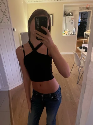 Svart croppad linnetopp med korsade band - Säljer en svart croppad linnetopp med smala axelband och snygga korsade band framtill. Toppen är ärmlös och har en tight passform som framhäver midjan. Perfekt till jeans eller kjol för en trendig look.